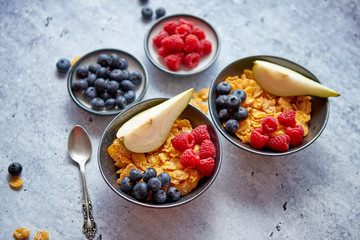 Healthy breakfast for two is served. Golden cornflakes with fresh fruits of raspberries, blueberries and pear in two ceramic bowls. Placed on stone background.