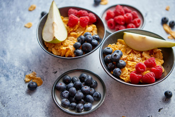 Healthy breakfast for two is served. Golden cornflakes with fresh fruits of raspberries, blueberries and pear in two ceramic bowls. Placed on stone background.