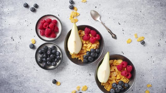 Healthy breakfast for two is served. Golden cornflakes with fresh fruits of raspberries, blueberries and pear in two ceramic bowls. Placed on stone background.