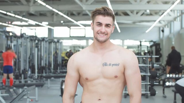Attractive shirtless blond male bodybuilder in shorts indoors in dark gym, showing muscular torso and ripped abs.