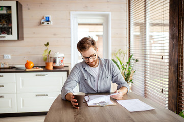 Young confident male examines house purchase agreement papers, buying the house, using smartphone...