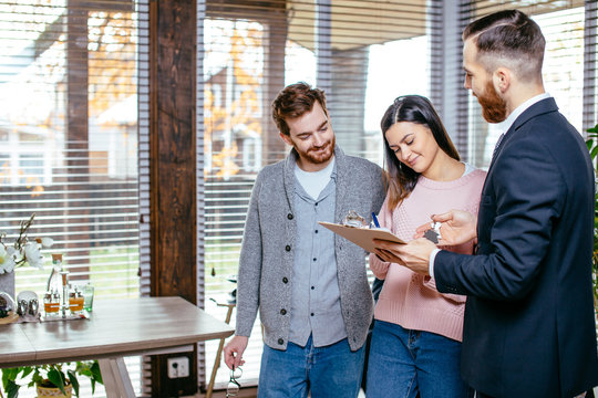 Smiling caucasian couple of attractive tenants shaking hands with landlord, receive house key, making rent deal