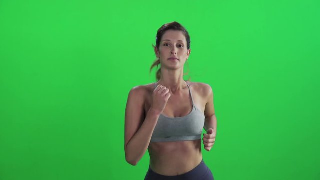 Young woman jogging in a medium frontal shot over a green screen, wearing a sports bra, looking into the lens, smiles.