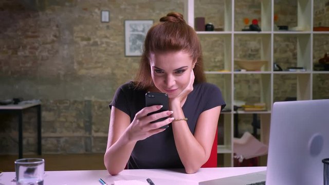 Cute caucasian business girl is scrolling her smartphone while sitting indoor and looking precisely at screen in office near laptop, relaxed, casual
