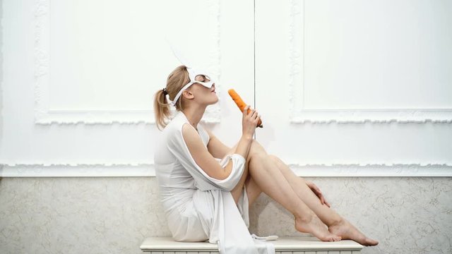 Girl dressed in a mask of a hare eating carrots on a white background. Brave woman in a mask eats carrots. Concept of modern fashion.