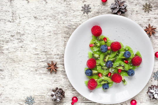 kiwi christmas tree with berries and coconut with pine cones over white wooden table. funny food idea for kids.