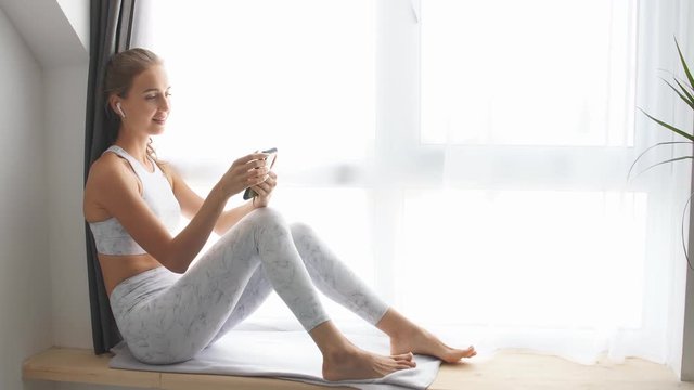 Woman sitting on window sill watching videos on smartphone and headphones.