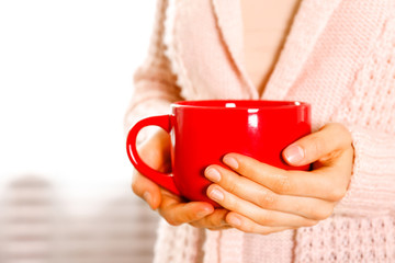Close up young woman's hands holding big cup of hot beverage. Female wearing fashionable oversized white knitted sweater, sitting home with mug of coffee. Background, copy space.