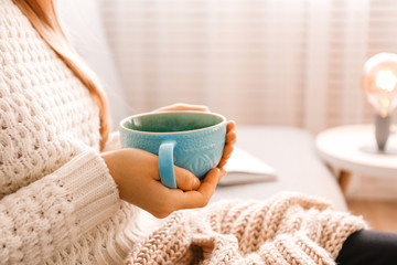 Close up young woman's hands holding big cup of hot beverage. Female wearing fashionable oversized white knitted sweater, sitting home with mug of coffee. Background, copy space.