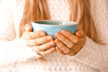 Close up young woman's hands holding big cup of hot beverage. Female wearing fashionable oversized white knitted sweater, sitting home with mug of coffee. Background, copy space.