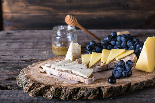 Assorted cheeses with grapes, bread, honey on dark wood background. Goat cheese with herbs. Natural wooden board. Italian appetizer. bruschetta with cheese. Still life of food. Breakfast concept.