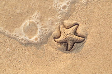 Starfish drawn on the beach sand being washed away by a wave. Foaming sea wave coming to wash a picture on wet yellow beach sand. Holiday and vocation message. View from above. Copy space.