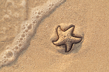 Fototapeta na wymiar Starfish drawn on the beach sand being washed away by a wave. Foaming sea wave coming to wash a picture on wet yellow beach sand. Holiday and vocation message. View from above. Copy space.