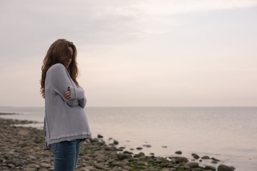 Young  Sad Pregnant Woman in a  Knitted Sweater Standing on a Deserted Beach. Sea on a Background.