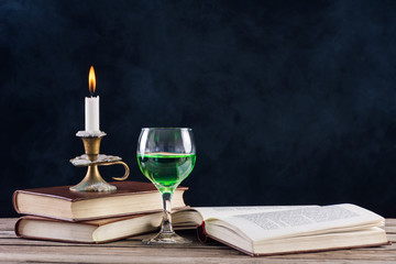 Absinthe drink in glass and books with candle in candlestick burning on retro wooden desk and smoky dark background. Green fairy inspiration and muse of writers and artists