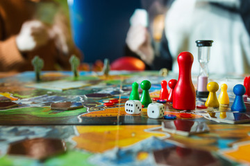 Board game concept- board game field, many figures, meeple, dice, coins and sandglass. Two people play holding cards on blurred background - obrazy, fototapety, plakaty