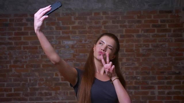 Cute caucasian female is taking selfie and showing her two fingers on her phone while standing on brick background, happy face and smiling person
