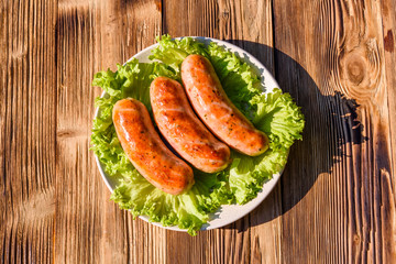 Ceramic plate with grilled sausages and lettuce leaves on wooden table. Top view