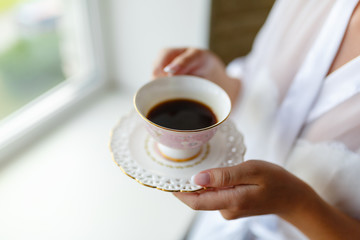 Girl in a Bathrobe sits on the windowsill next to the window and drinks coffee from a mug with a saucer. Young woman in a bathrobe drinking hot espresso or cappuccino in the morning on the windowsill.