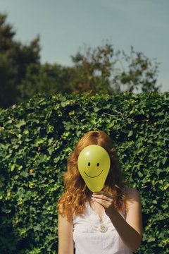 Young redheaded woman holds a yellow balloon with a smiley face in front of her own face.