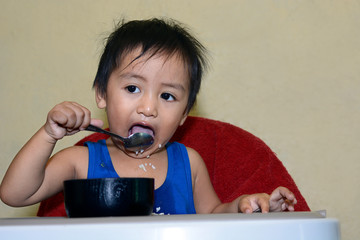 One 1 year old Asian baby boy learning to eat by himself by spoon, messy on baby dining chair at home