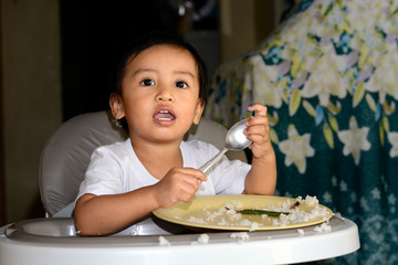 One 1 year old Asian baby boy learning to eat by himself, messy on baby dining chair at home