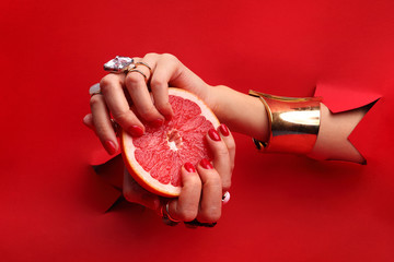 Manicure. Kobiece dłonie z  czerwonymi paznokciami przez otwór w czerwonym tle  trzymają owoc grejpfruta. - obrazy, fototapety, plakaty