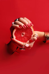 Kobiece eleganckie dłonie. Kobiece dłonie z  czerwonymi paznokciami przez otwór w czerwonym tle  trzymają owoc grejpfruta. - obrazy, fototapety, plakaty