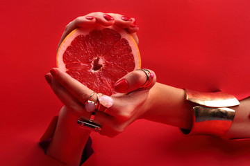 Kobiece eleganckie dłonie. Kobiece dłonie z  czerwonymi paznokciami przez otwór w czerwonym tle  trzymają owoc grejpfruta. - obrazy, fototapety, plakaty