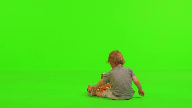 A toddler sits and plays with a popcorn bawl and some toys, full over the shoulder shot on a green screen.
