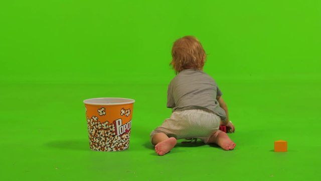 A toddler sits and put some toys inside a popcorn bawl over a green screen.