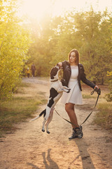 Fototapeta premium Cute young girl and dog walk in autumn park. Dog is spotted, hunting, fold and short-legged. Girl in white dress and black leather jacket. Pet is jumping for treat.