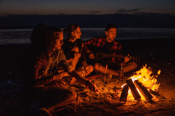 Camp on the beach. Group of young friends having picnic with bonfire. Man is playing guitar