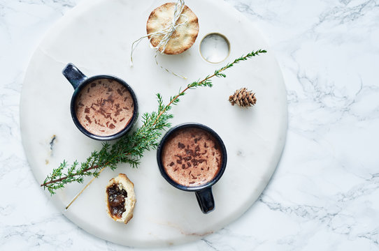 Two mugs of hot chocolate and Christmas mince pies on marble board.