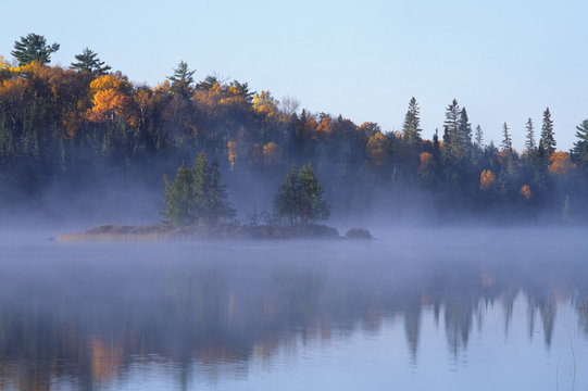Fog and mist arising from a lake in Quetico Provincial Park, Ontario, Canada in late autumn.  Gold leaf trees rimming the shoreline, a small island sitting out in the fog.  Photographed on Fuji Velvia film.