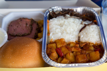 Food served in a passenger aircraft during the flight. Meal on the tray. Salad, meat appetizer, bread bun. Hot dish in the aluminum lunch box: chicken, rice and vegetables.