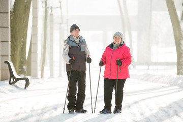 Senior couple walking with nordic walking poles in winter park. Mature woman and old man doing exercise outdoors. Healthy lifestyle concept.