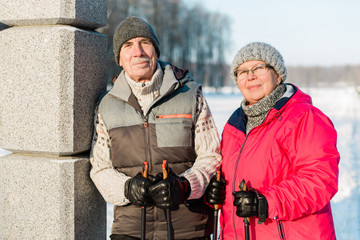 Pretty senior couple standing with nordic walking poles in winter park. Mature woman and old man resting outdoors. Healthy lifestyle concept.