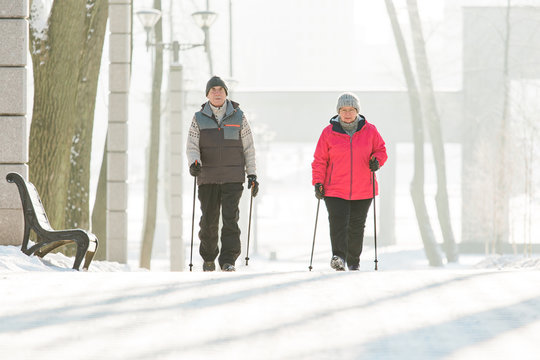 Senior Couple Walking With Nordic Walking Poles In Winter Park. Mature Woman And Old Man Doing Exercise Outdoors. Healthy Lifestyle Concept.