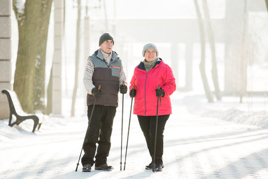 Senior Couple Walking With Nordic Walking Poles In Winter Park. Mature Woman And Old Man Doing Exercise Outdoors. Healthy Lifestyle Concept.