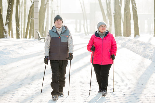 Senior Couple Walking With Nordic Walking Poles In Winter Park. Mature Woman And Old Man Doing Exercise Outdoors. Healthy Lifestyle Concept.