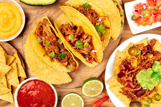 A photo of Mexican food, including tacos, guacamole, pico de gallo, and nachos with chili con carne, shot from the top with ingredients on a wooden background