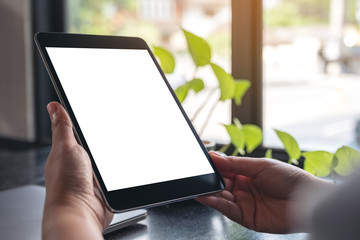 Mockup image of hands holding black tablet pc with blank white desktop screen with green leaves on wooden table