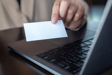 A hand holding and giving white empty business card with laptop on wooden table in office