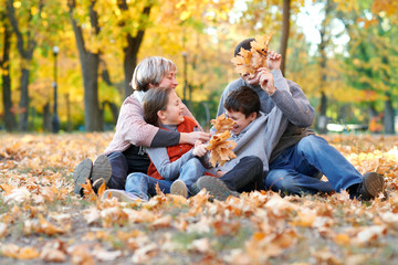 Happy family sit in autumn city park on fallen leaves. Children and parents posing, smiling, playing and having fun. Bright yellow trees.