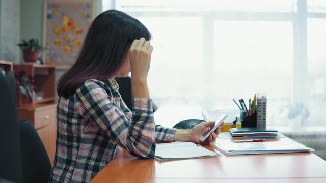 Side view, young female teacher dressed in checkered shirt sits at the table in the classroom, she is looking for information using the browser on the smartphone.