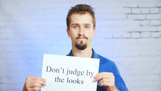 Person hold the Don't judge by the looks sign 4K. Static portrait shot of man in blue elegant shirt in focus hold a sign with words. Background wall with a blue light spill.