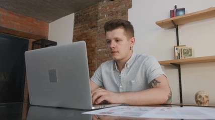 Attractive smiling Caucasian young man with cool tattoos sitting at table and finishing work. Glad stunning guy closing laptop. Successful project. Indoors.
