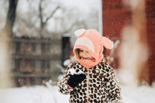 Toddler girl in cute cowl hat looking at snow before eating it.
