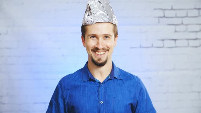 Person with tin-foil hat smiling into camera 4K. Static portrait shot of a man in focus wearing a blue elegant shirt and a tin-foil hat, smiling into the camera.